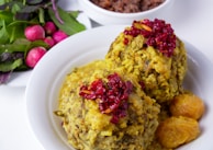 Two large servings of a rice and herb dish topped with red berries are presented on a white plate. The dish is accompanied by small yellow cooked fruits or vegetables. There is a side of fresh greens and radishes, and a bowl with a dark bean mixture is partially visible in the background.