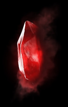 A vibrant red crystal, partially translucent with light reflecting on its facets, is suspended against a dark background. The surrounding area has a misty glow, enhancing the mysterious and enchanted appearance.