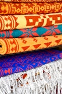 A collection of intricately patterned, colorful woven rugs is neatly stacked. The rugs feature bold geometric designs with dominant colors including orange, red, and blue. The top rugs are rolled while the bottom ones display fringes on their edges.