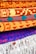 A collection of intricately patterned, colorful woven rugs is neatly stacked. The rugs feature bold geometric designs with dominant colors including orange, red, and blue. The top rugs are rolled while the bottom ones display fringes on their edges.