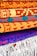 A collection of intricately patterned, colorful woven rugs is neatly stacked. The rugs feature bold geometric designs with dominant colors including orange, red, and blue. The top rugs are rolled while the bottom ones display fringes on their edges.