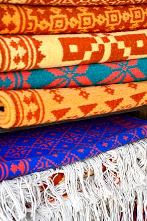 A collection of intricately patterned, colorful woven rugs is neatly stacked. The rugs feature bold geometric designs with dominant colors including orange, red, and blue. The top rugs are rolled while the bottom ones display fringes on their edges.