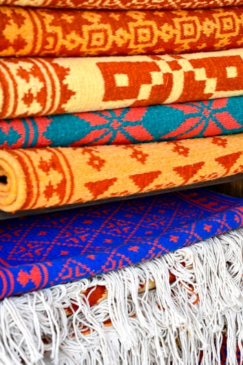 A collection of intricately patterned, colorful woven rugs is neatly stacked. The rugs feature bold geometric designs with dominant colors including orange, red, and blue. The top rugs are rolled while the bottom ones display fringes on their edges.