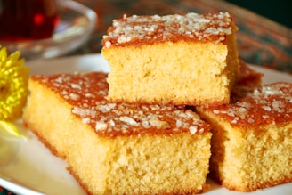 Three pieces of golden, fluffy cake are stacked on a white plate, topped with granulated sugar or coarse salt, giving a textured surface. A blurred background includes a hint of a yellow flower and possibly a cup.