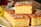 Three pieces of golden, fluffy cake are stacked on a white plate, topped with granulated sugar or coarse salt, giving a textured surface. A blurred background includes a hint of a yellow flower and possibly a cup.