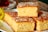 Three pieces of golden, fluffy cake are stacked on a white plate, topped with granulated sugar or coarse salt, giving a textured surface. A blurred background includes a hint of a yellow flower and possibly a cup.