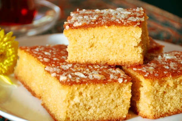 Three pieces of golden, fluffy cake are stacked on a white plate, topped with granulated sugar or coarse salt, giving a textured surface. A blurred background includes a hint of a yellow flower and possibly a cup.