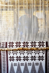 A hand-woven textile with geometric patterns featuring stars and trees is displayed on a loom. The loom threads are tightly strung vertically, and a person is partially visible behind them, working on the weaving.