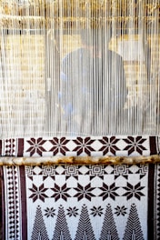 A hand-woven textile with geometric patterns featuring stars and trees is displayed on a loom. The loom threads are tightly strung vertically, and a person is partially visible behind them, working on the weaving.