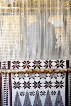 A hand-woven textile with geometric patterns featuring stars and trees is displayed on a loom. The loom threads are tightly strung vertically, and a person is partially visible behind them, working on the weaving.