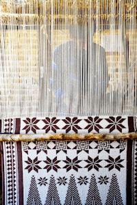 A hand-woven textile with geometric patterns featuring stars and trees is displayed on a loom. The loom threads are tightly strung vertically, and a person is partially visible behind them, working on the weaving.
