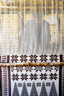 A hand-woven textile with geometric patterns featuring stars and trees is displayed on a loom. The loom threads are tightly strung vertically, and a person is partially visible behind them, working on the weaving.
