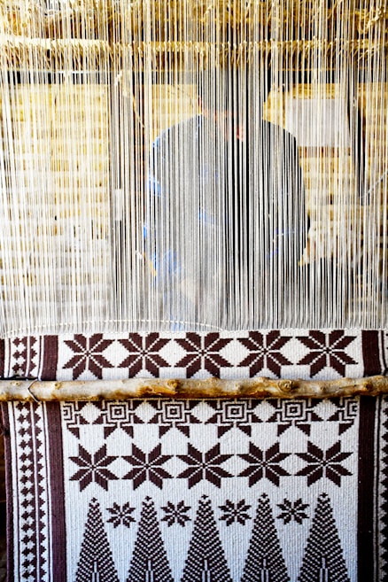 A hand-woven textile with geometric patterns featuring stars and trees is displayed on a loom. The loom threads are tightly strung vertically, and a person is partially visible behind them, working on the weaving.