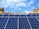 Rows of blue solar panels are arranged on a rooftop under a bright blue sky. In the background, a variety of architectural buildings with different styles and shapes are visible.