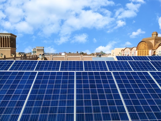 Rows of blue solar panels are arranged on a rooftop under a bright blue sky. In the background, a variety of architectural buildings with different styles and shapes are visible.