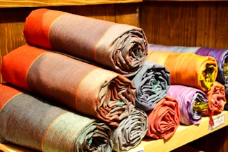 A collection of rolled-up fabric pieces neatly stacked on wooden shelves. The fabrics feature various colors, including reds, oranges, purples, and browns, each with a subtle checkered pattern. The texture of the woven fabric is visible, and the arrangement exudes an organized and inviting appearance.