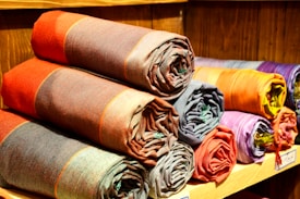 A collection of rolled-up fabric pieces neatly stacked on wooden shelves. The fabrics feature various colors, including reds, oranges, purples, and browns, each with a subtle checkered pattern. The texture of the woven fabric is visible, and the arrangement exudes an organized and inviting appearance.