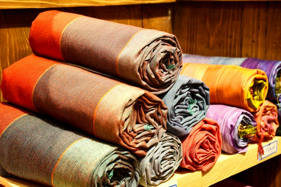 A collection of rolled-up fabric pieces neatly stacked on wooden shelves. The fabrics feature various colors, including reds, oranges, purples, and browns, each with a subtle checkered pattern. The texture of the woven fabric is visible, and the arrangement exudes an organized and inviting appearance.