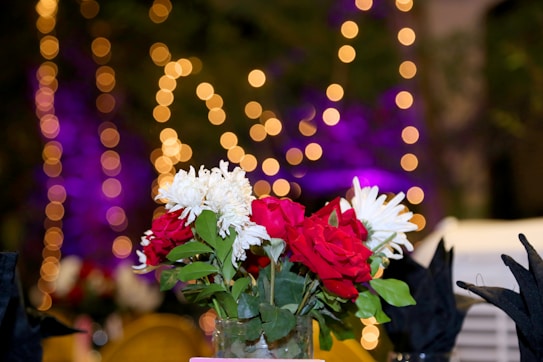 A bouquet of red and white flowers is the focal point, with a blurred background featuring warm, circular lights creating a bokeh effect. The setting is vibrant with purples and yellows enhancing the festive atmosphere.