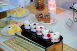 A variety of dessert cups are elegantly arranged on a glass and wooden stand. The front row features cups with layers of whipped cream and strawberries, topped with a cherry. In the background, there are cups of yellow mousse and chocolate desserts decorated with cream and toppings, all set on a decorative table with a golden patterned mat and ambient lighting.