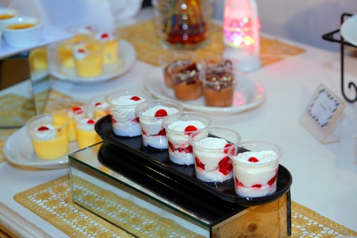 A variety of dessert cups are elegantly arranged on a glass and wooden stand. The front row features cups with layers of whipped cream and strawberries, topped with a cherry. In the background, there are cups of yellow mousse and chocolate desserts decorated with cream and toppings, all set on a decorative table with a golden patterned mat and ambient lighting.