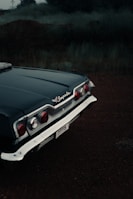 The back end of a classic Chevrolet Impala, showcasing the car's distinctive tail light design and chrome detailing, is parked on a dark, dirt ground with a hazy backdrop of overgrown grass and trees. The photograph exudes a vintage atmosphere with muted lighting.