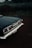 The back end of a classic Chevrolet Impala, showcasing the car's distinctive tail light design and chrome detailing, is parked on a dark, dirt ground with a hazy backdrop of overgrown grass and trees. The photograph exudes a vintage atmosphere with muted lighting.