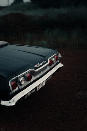 The back end of a classic Chevrolet Impala, showcasing the car's distinctive tail light design and chrome detailing, is parked on a dark, dirt ground with a hazy backdrop of overgrown grass and trees. The photograph exudes a vintage atmosphere with muted lighting.