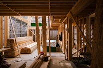 A partially constructed wooden building featuring an array of wooden beams and planks neatly stacked and assembled. Scaffolding is visible on one side, with various construction tools and equipment scattered around the interior. Natural light filters through the unfinished structure, creating a warm atmosphere.