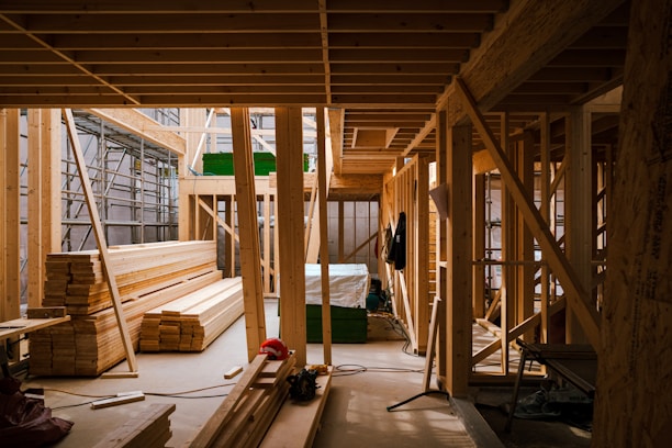 A partially constructed wooden building featuring an array of wooden beams and planks neatly stacked and assembled. Scaffolding is visible on one side, with various construction tools and equipment scattered around the interior. Natural light filters through the unfinished structure, creating a warm atmosphere.