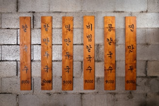 Wooden planks with Korean text are mounted on a gray brick wall. Each plank has black lettering and numbers indicating prices. The planks vary slightly in width and height.