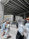 A group of people dressed in white clothing, some wearing head coverings, are walking under a large, modern overpass structure. Several individuals are holding umbrellas to shield themselves from the sun. The scene appears to be a pilgrimage, with people carrying bags and looking purposeful.