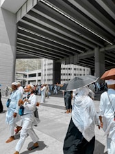 A group of people dressed in white clothing, some wearing head coverings, are walking under a large, modern overpass structure. Several individuals are holding umbrellas to shield themselves from the sun. The scene appears to be a pilgrimage, with people carrying bags and looking purposeful.