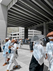 A group of people dressed in white clothing, some wearing head coverings, are walking under a large, modern overpass structure. Several individuals are holding umbrellas to shield themselves from the sun. The scene appears to be a pilgrimage, with people carrying bags and looking purposeful.