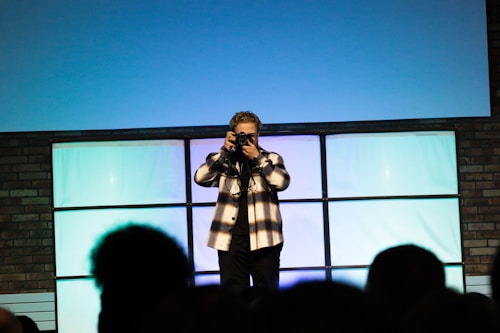 A person is standing indoors holding a camera, facing the audience. The background consists of a tiled wall divided into squares with a soft gradient of blue and green lighting. The audience is visible in the foreground as silhouettes, creating a contrast with the brightly lit background.