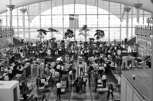 A busy airport security checkpoint with numerous people in line and security personnel conducting checks. There are various security machines and equipment in operation. The space is large with high ceilings, expansive windows, and an American flag hanging in the background. Potted trees can be seen near the windows, adding a touch of greenery to the scene. The area is well-lit with natural light streaming in through the windows.