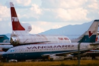 A scene of two airplanes, one with 'TWA' and 'Trans World' markings, and another with 'Vanguard' livery. The aircraft are parked on a field, and the surrounding landscape includes mountains under a partly cloudy sky.
