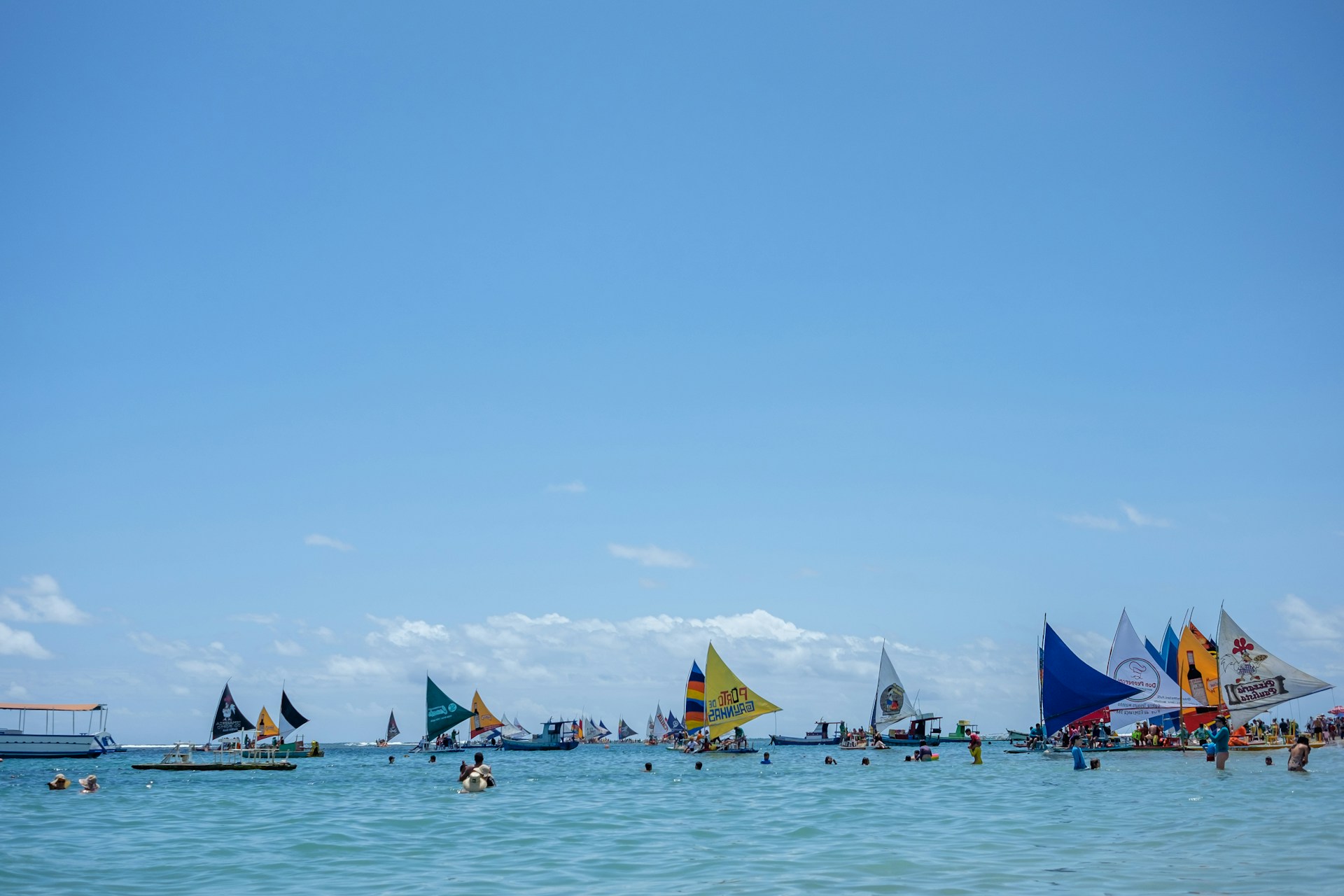 A bright, sunny day in Didim with colorful sailboats gliding over the clear blue Aegean Sea near the coastline.