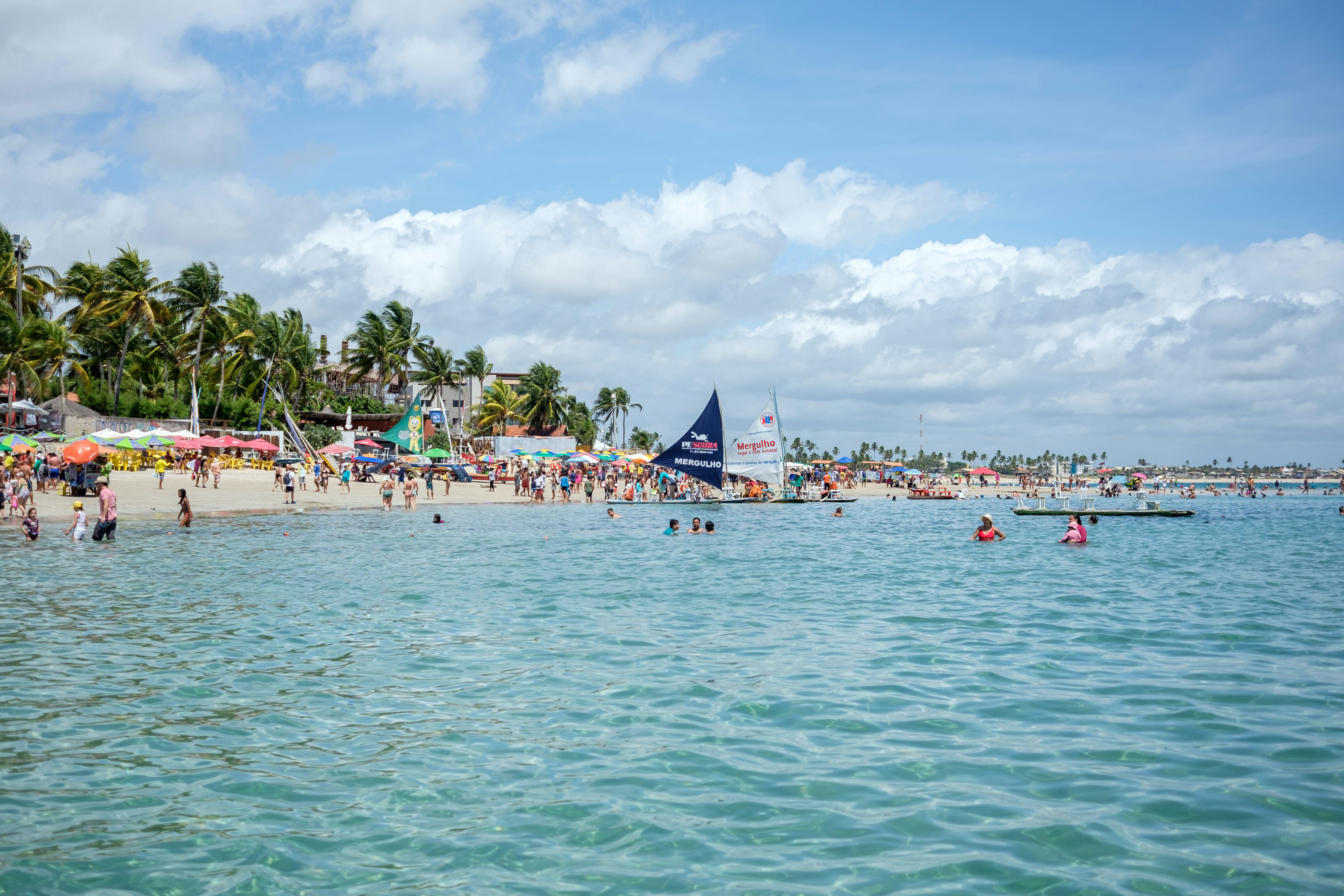 Beach with crystal clear water lined with sail boats and packed with people in Porto de Galinhas