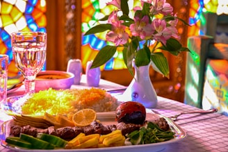 A beautifully set dining table with a plate of grilled meat skewers, assorted vegetables, and a slice of lemon. A side of saffron rice is served, next to a glass of water and a bowl of soup. There is a small vase with pink flowers. The background is brightly lit with colorful stained glass patterns, creating a vibrant ambiance.