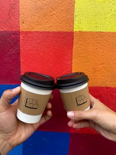 Two hands holding disposable coffee cups with brown sleeves against a vibrant, multicolored geometric background.