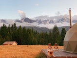 A serene landscape featuring a grassy field with a small wooden cabin, set against a backdrop of lush green trees and majestic snow-capped mountains under a clear sky. Two wooden chairs and a small table are placed on a wooden deck beside a dome-shaped structure, suggesting a peaceful retreat in nature.