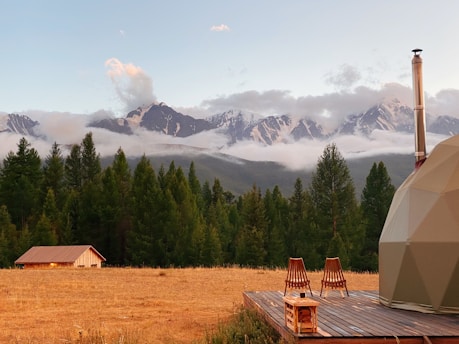 A serene landscape featuring a grassy field with a small wooden cabin, set against a backdrop of lush green trees and majestic snow-capped mountains under a clear sky. Two wooden chairs and a small table are placed on a wooden deck beside a dome-shaped structure, suggesting a peaceful retreat in nature.