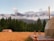 A serene landscape featuring a grassy field with a small wooden cabin, set against a backdrop of lush green trees and majestic snow-capped mountains under a clear sky. Two wooden chairs and a small table are placed on a wooden deck beside a dome-shaped structure, suggesting a peaceful retreat in nature.