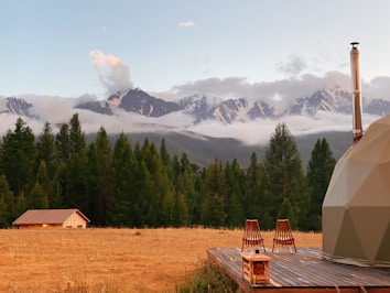 A serene landscape featuring a grassy field with a small wooden cabin, set against a backdrop of lush green trees and majestic snow-capped mountains under a clear sky. Two wooden chairs and a small table are placed on a wooden deck beside a dome-shaped structure, suggesting a peaceful retreat in nature.