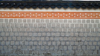 A traditional stone wall featuring a layer of clay bricks highlighted with orange and white colors. The top of the wall displays a series of dark gray, rounded roof tiles, which add a distinct architectural element.