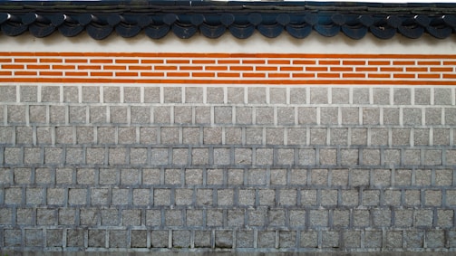 A traditional stone wall featuring a layer of clay bricks highlighted with orange and white colors. The top of the wall displays a series of dark gray, rounded roof tiles, which add a distinct architectural element.