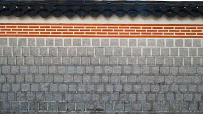 A traditional stone wall featuring a layer of clay bricks highlighted with orange and white colors. The top of the wall displays a series of dark gray, rounded roof tiles, which add a distinct architectural element.