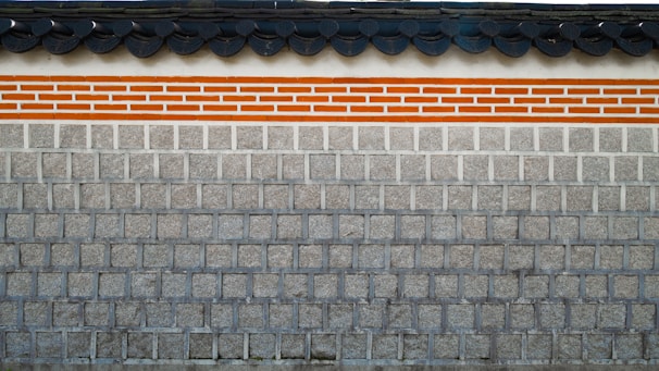 A traditional stone wall featuring a layer of clay bricks highlighted with orange and white colors. The top of the wall displays a series of dark gray, rounded roof tiles, which add a distinct architectural element.