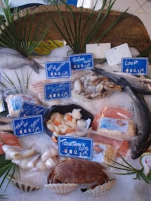 A display of various seafood items on ice, including whole fish, shellfish, and packaged items, with price tags in French. The arrangement is decorated with green palm-like leaves.